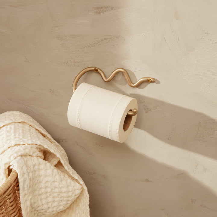 Curvature toiletpapirholder fra ferm Living på en lys stenmur ved siden af en rottingkurv